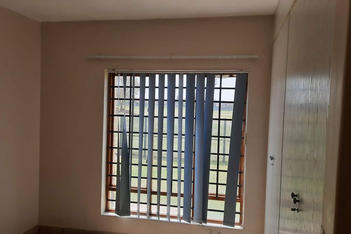 A vacant bedroom featuring built-in cupboards, security bars on the window, and vertical blinds in need of repair.