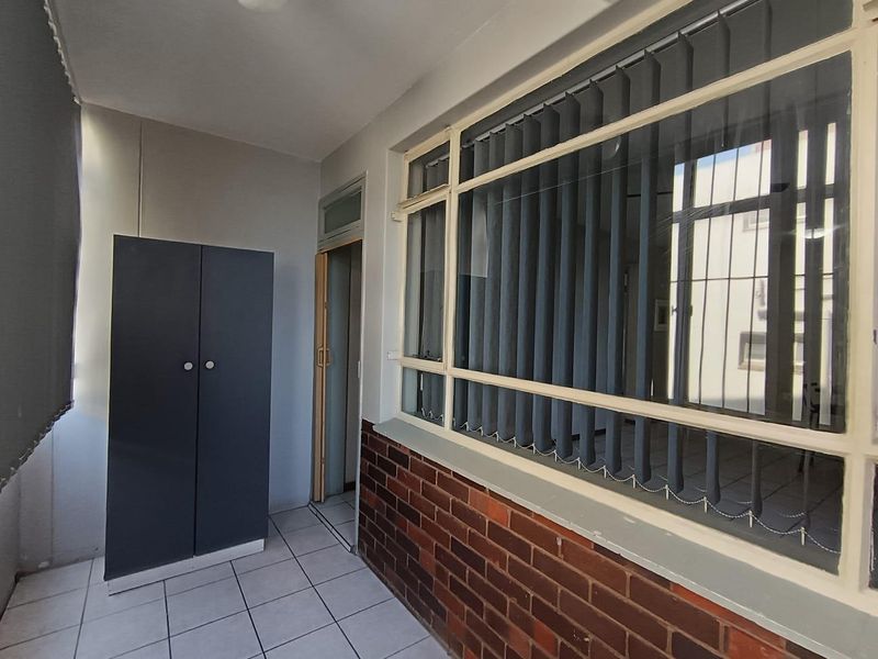 A covered balcony or enclosed patio area featuring tiled flooring, brick wall detailing, and large windows with vertical blinds.