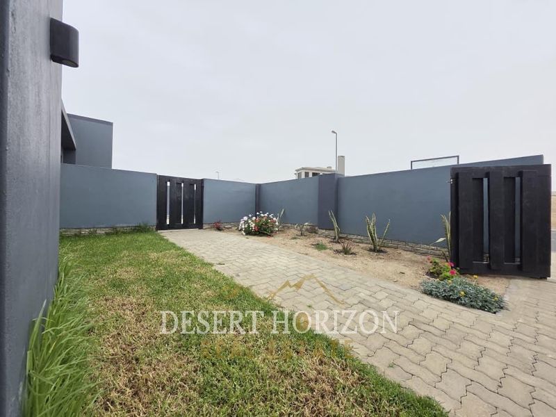 A front entrance area featuring a paved walkway, dark grey boundary walls, and timber gates, providing a clean and modern street-facing access point.