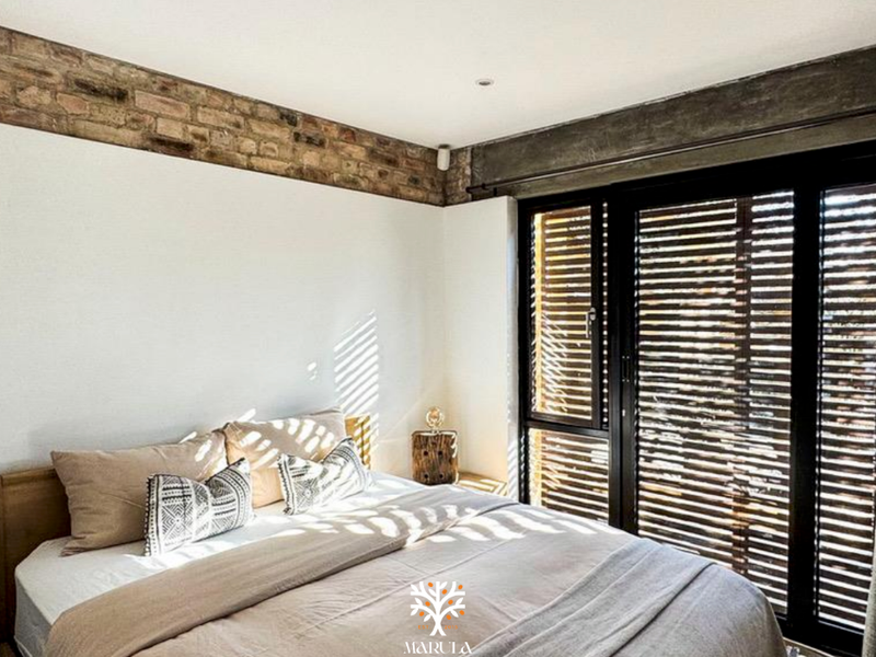 A modern bedroom featuring exposed brick and concrete architectural details, complemented by black-framed sliding doors with integrated timber shutters for natural light control.