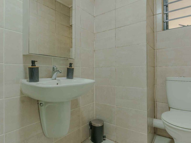 A functional bathroom featuring a pedestal washbasin, toilet, and full-height wall tiling, complemented by a built-in medicine cabinet and window security bars.