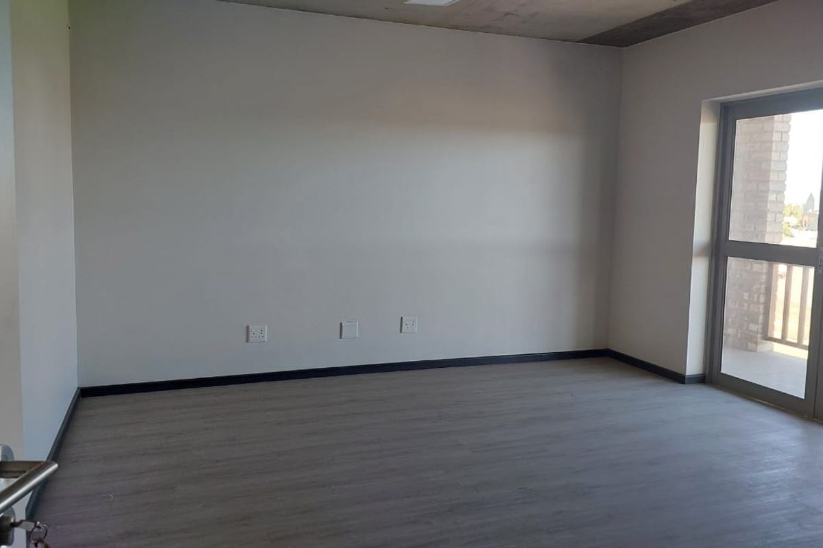 A bright, empty bedroom featuring modern grey laminate flooring, neutral walls, and a glass door providing access to an outdoor balcony.