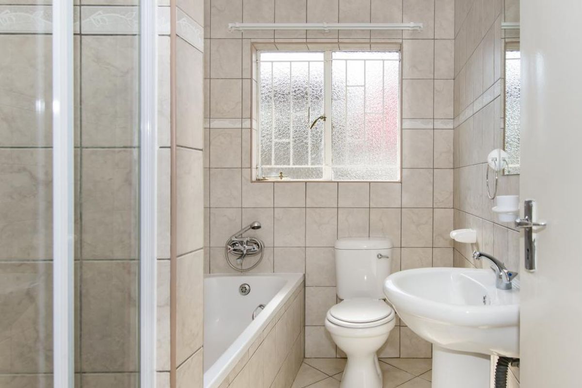 A functional bathroom featuring a bathtub, pedestal basin, and toilet, finished with neutral wall and floor tiling and a security-grilled window.