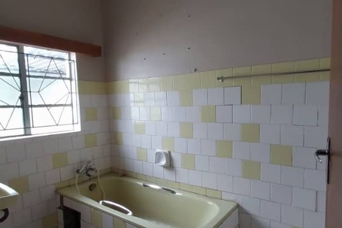 A dated bathroom featuring a built-in bathtub with tiled surround, ceramic floor tiles, and a security-barred window.