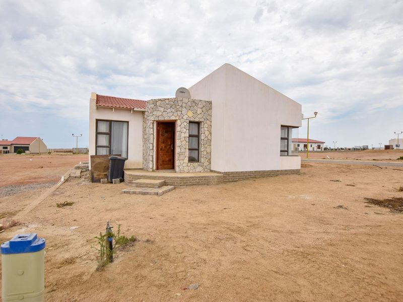 Front elevation of a detached house featuring stone cladding detail, tiled roof, and aluminium window frames, set on an undeveloped sandy plot.