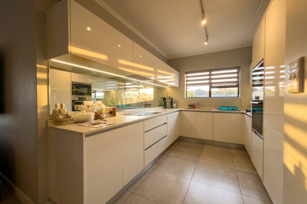 Modern kitchen featuring high-gloss cabinetry, integrated oven, sleek stone countertops, and a mirrored splashback, illuminated by contemporary track lighting.
