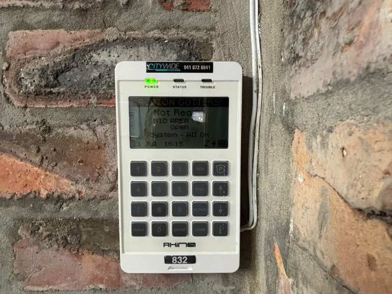 A wall-mounted security alarm keypad installed on an exposed brick wall surface.