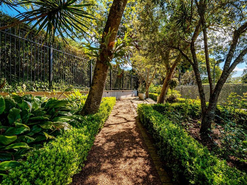 A well-maintained garden featuring a gravel pathway lined with manicured hedges, mature trees, and secure metal perimeter fencing.