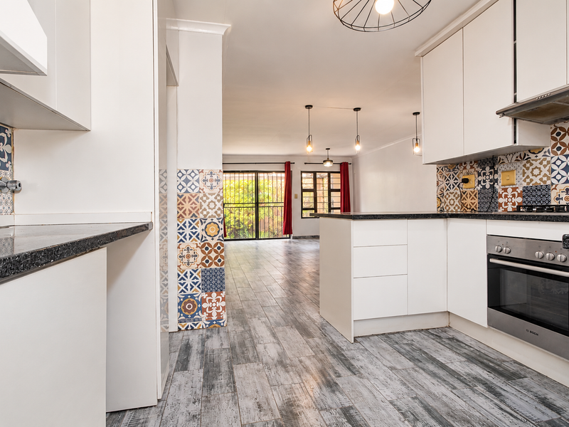 Modern open-plan kitchen featuring white cabinetry, granite countertops, and decorative tile backsplash, flowing into a spacious living area with wood-look tile flooring.