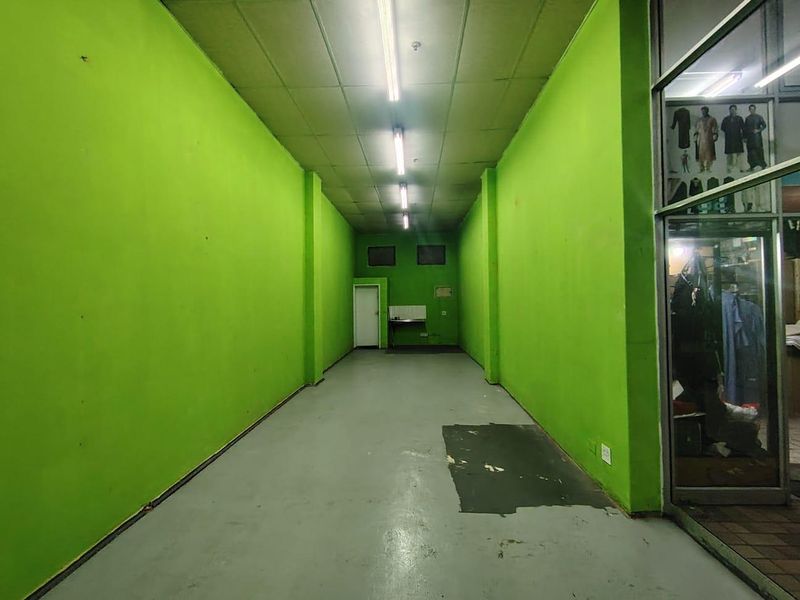 An empty retail utility space featuring concrete flooring, suspended ceiling with fluorescent lighting, and a glass storefront entrance.