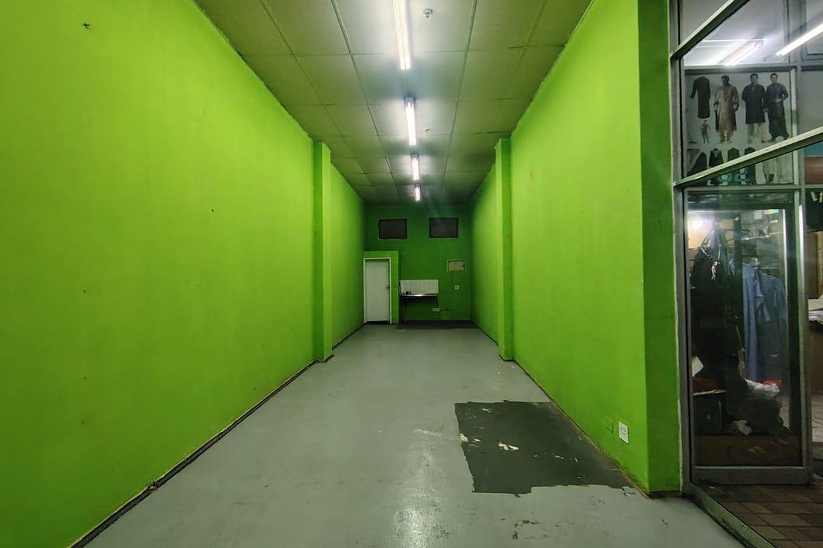 An empty retail utility space featuring concrete flooring, suspended ceiling with fluorescent lighting, and a glass storefront entrance.