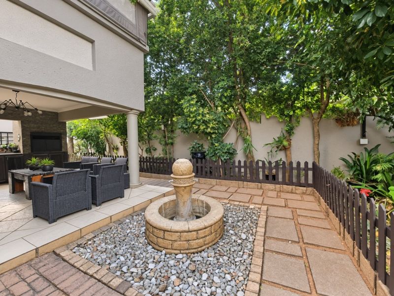 A well-maintained covered patio area featuring a stone fountain, paved flooring, and a built-in braai, set within a landscaped garden environment.