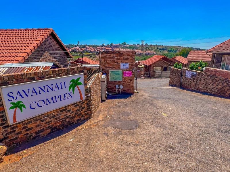 Entrance view of Savannah Complex featuring a brick boundary wall, paved driveway, and residential townhouse units with tiled roofs.