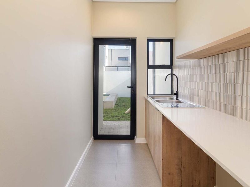 A modern, minimalist scullery featuring sleek timber cabinetry, stone countertops, a stainless steel sink, and direct access to the rear garden.