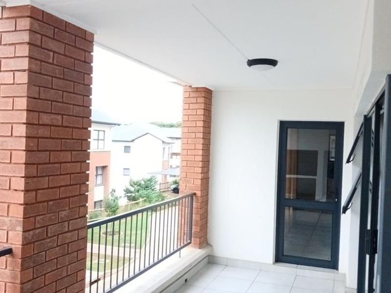 A clean, covered balcony featuring tiled flooring, brick support pillars, and a metal balustrade, offering a view of the surrounding residential complex.