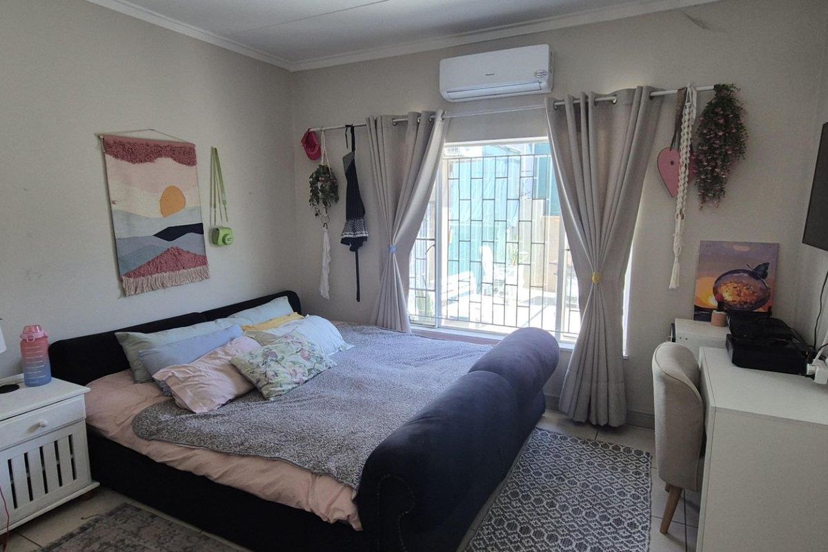 A functional bedroom featuring tiled flooring, a wall-mounted split-system air conditioner, and a window fitted with security bars.