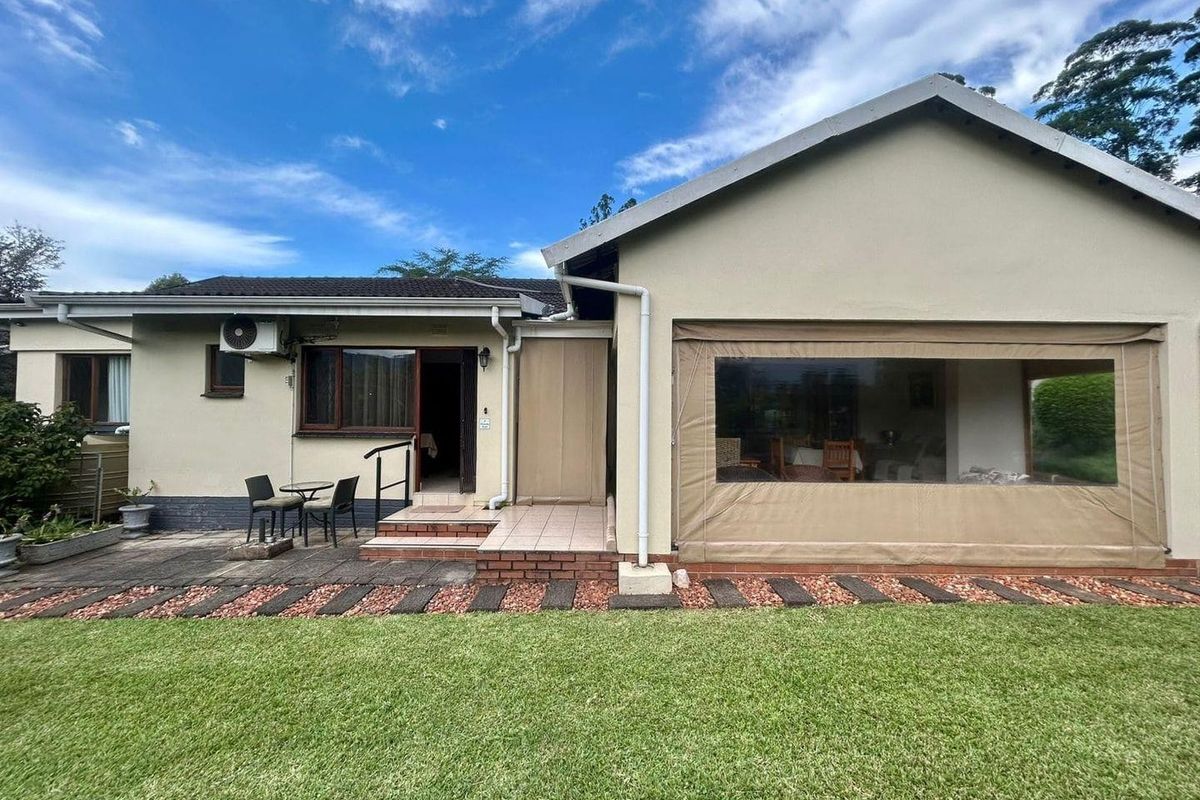 Rear view of the house featuring a paved patio area, a covered outdoor space with roll-down blinds, and a well-maintained lawn.
