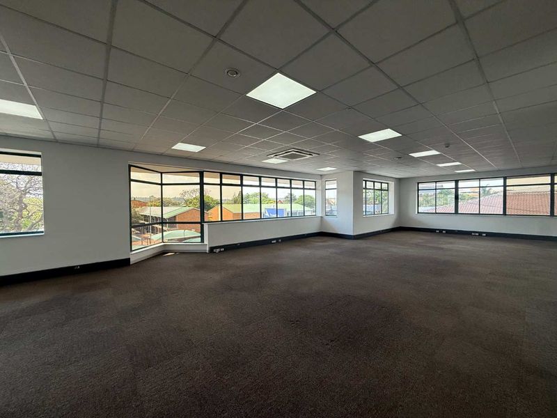 Spacious, open-plan office area featuring large windows, suspended ceilings with recessed lighting, and carpeted flooring, offering a bright and professional workspace.