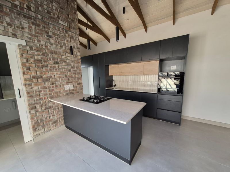 Modern kitchen featuring a central island with gas hob, sleek cabinetry, stone countertops, and a striking exposed brick wall under a vaulted timber ceiling.