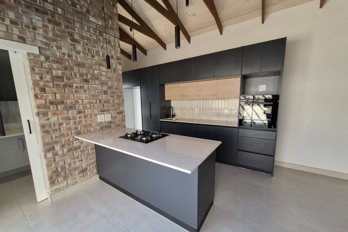 Modern kitchen featuring a central island with gas hob, sleek cabinetry, stone countertops, and a striking exposed brick wall under a vaulted timber ceiling.