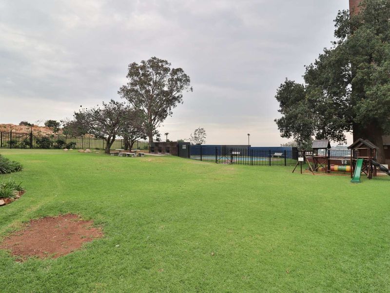 Spacious communal garden area featuring a lawn, built-in braai facilities, a fenced swimming pool, and a children's playground.