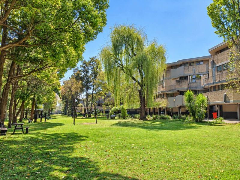 A well-maintained communal garden area featuring expansive green lawns and mature trees, adjacent to the brick apartment building complex.