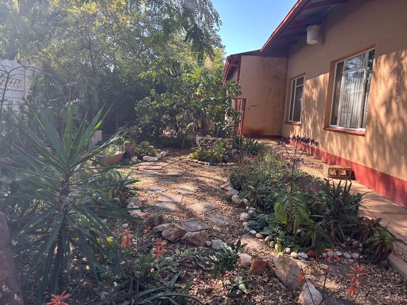 A rear garden area featuring a stone pathway, established landscaping, and a view of the house exterior with metal-framed windows.