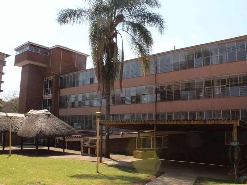 Rear view of a multi-story apartment building featuring brick construction, louvered windows, a communal thatched lapa area, and a covered walkway.