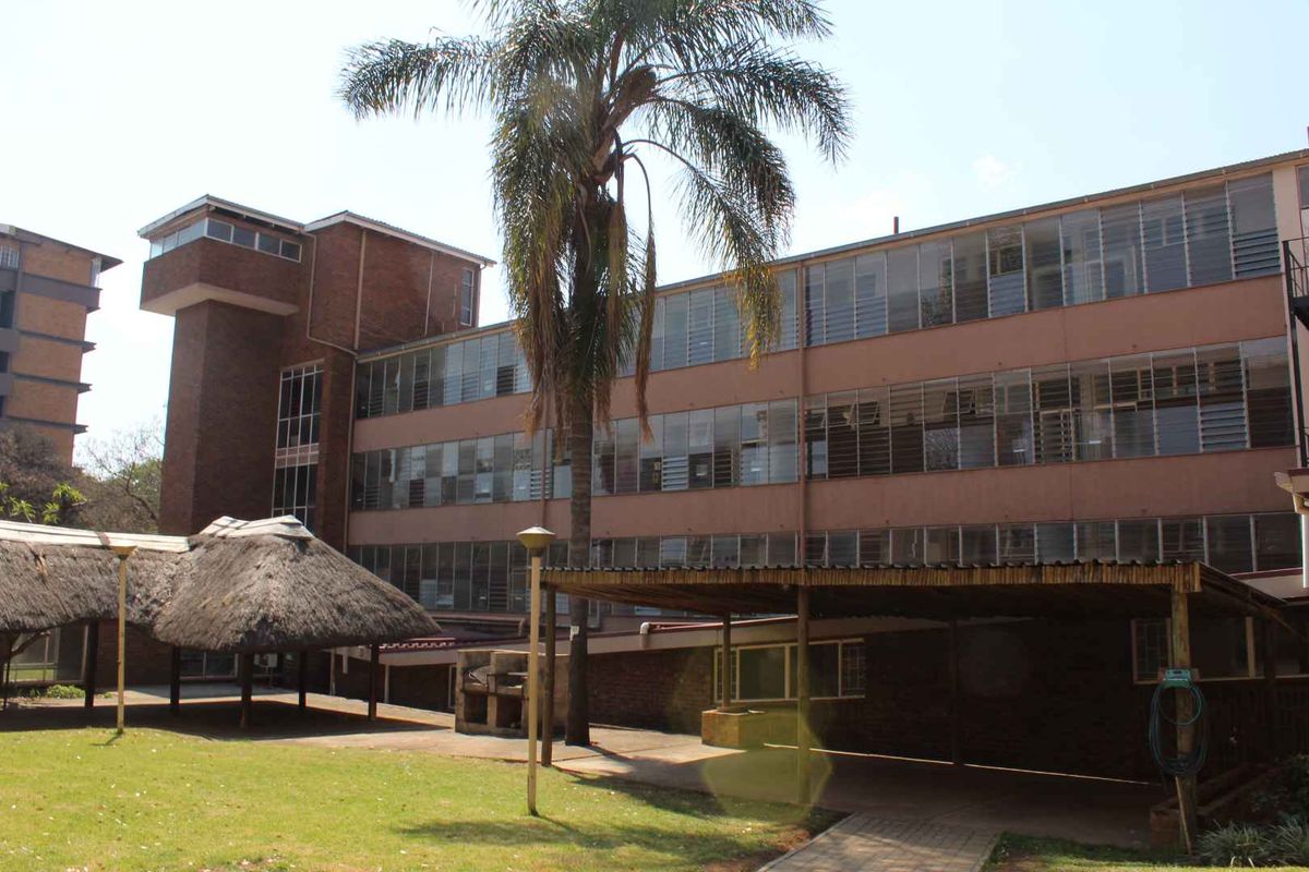 Rear view of a multi-story apartment building featuring brick construction, louvered windows, a communal thatched lapa area, and a covered walkway.