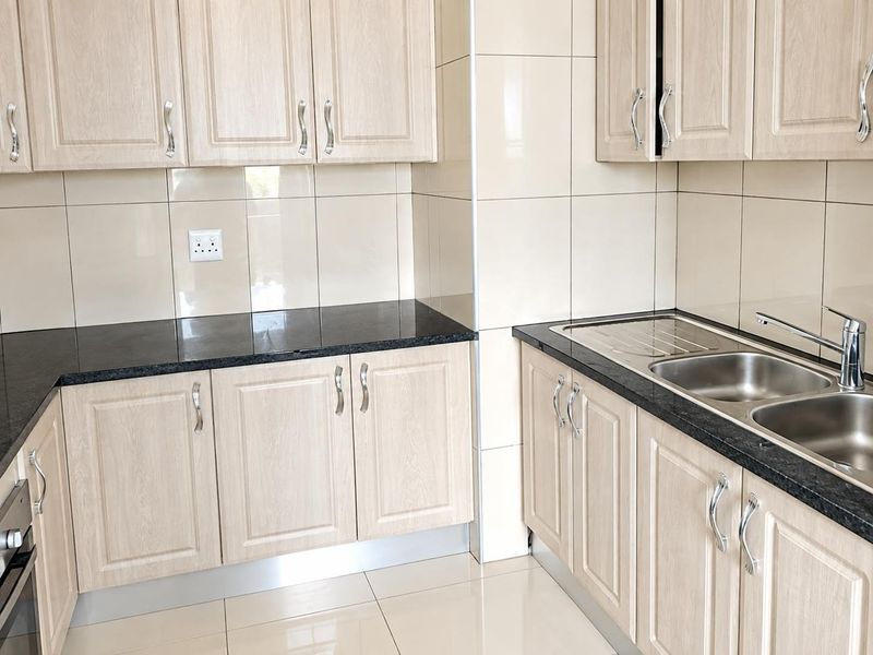 A clean, modern kitchen featuring light wood-finish cabinetry, dark granite countertops, double stainless steel sink, and neutral ceramic tiling throughout.
