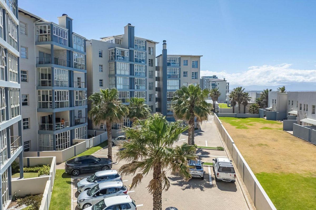 Elevated view of the apartment complex exterior, featuring multi-story residential blocks, paved parking area, and landscaped grounds with palm trees.