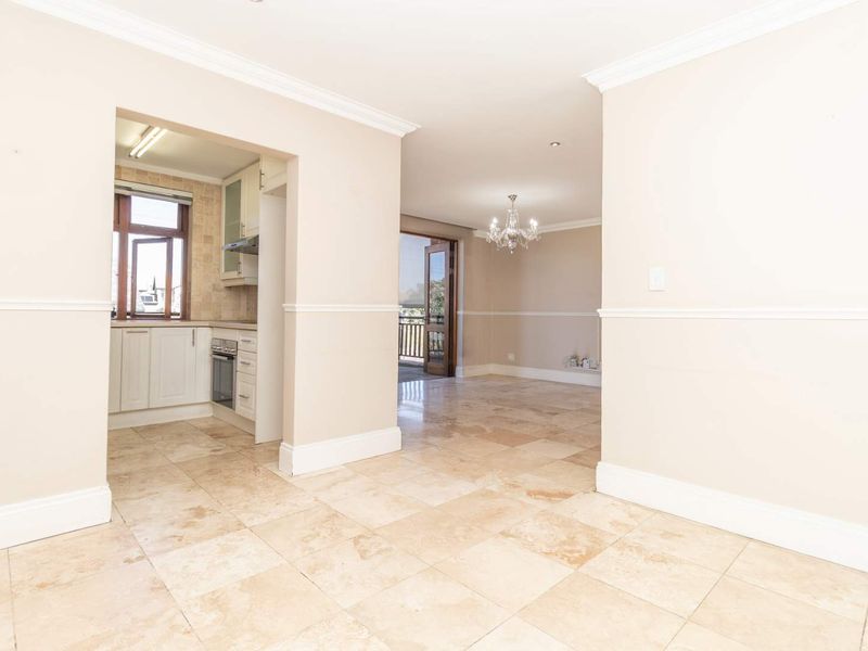 Spacious open-plan living area featuring travertine flooring, crown molding, and a functional kitchen with built-in cabinetry and appliances, leading to an outdoor balcony.