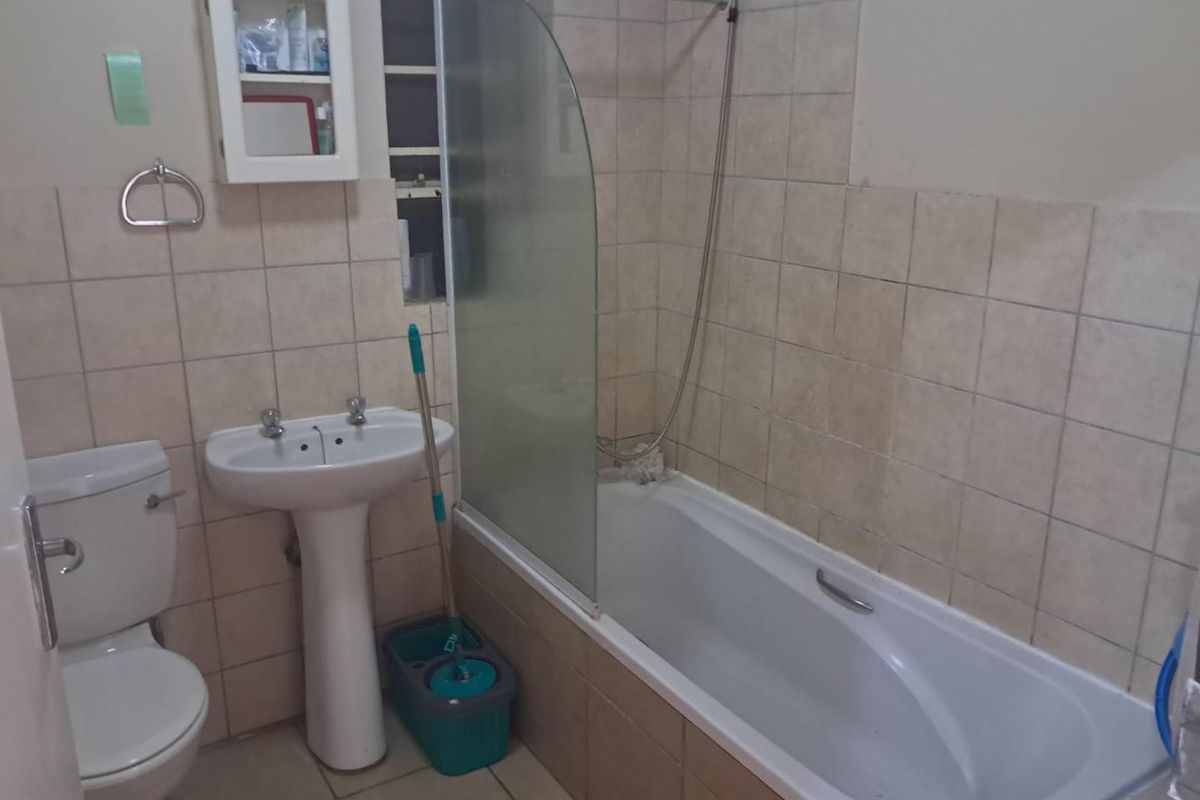 A functional bathroom featuring a pedestal basin, toilet, and a bathtub with a glass shower screen, finished with neutral wall and floor tiles.
