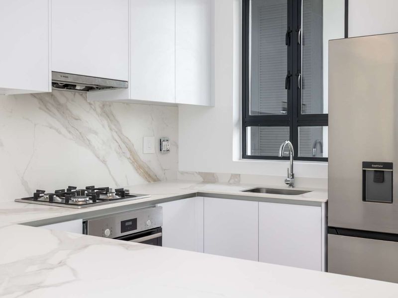 Modern kitchen featuring white cabinetry, marble-look stone countertops and backsplash, built-in gas hob, oven, and stainless steel sink with a window view.