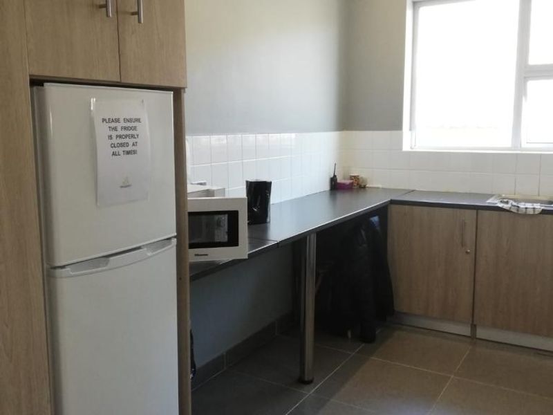 A functional office kitchenette featuring light wood-tone cabinetry, dark countertops, tiled backsplash, and tiled flooring with integrated sink and lighting.
