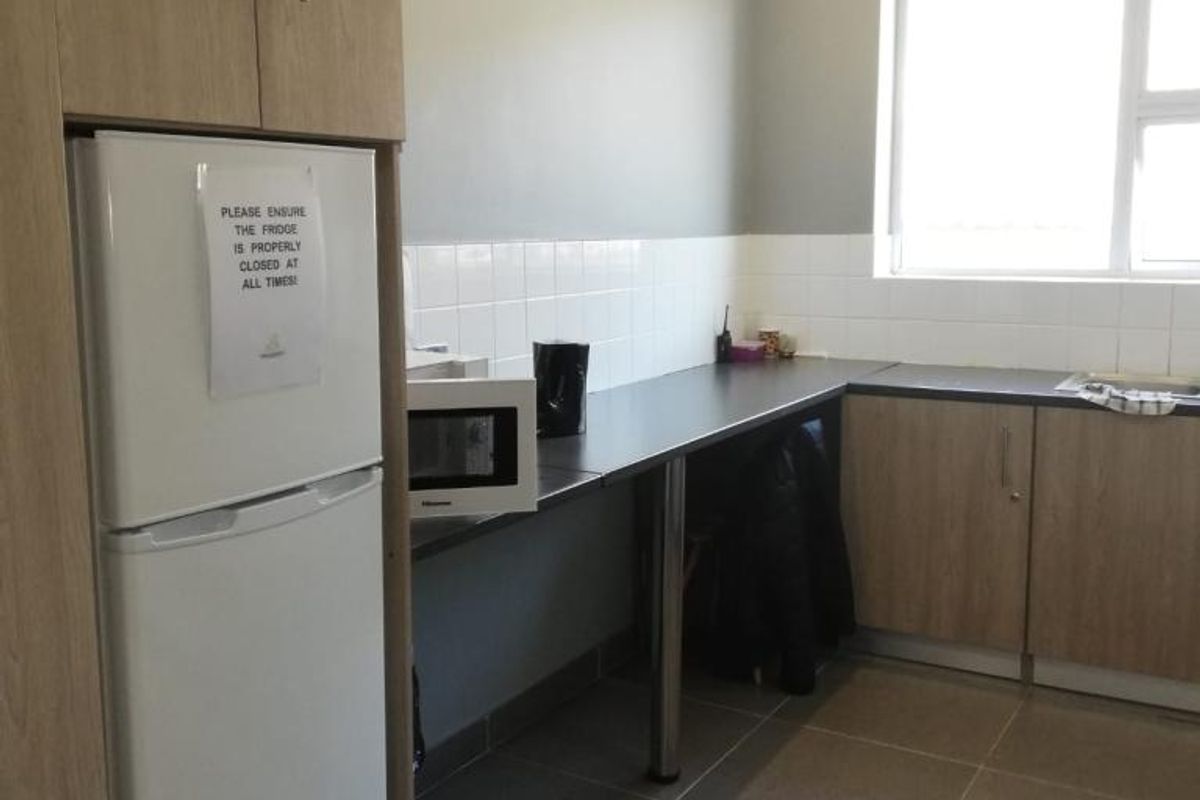 A functional office kitchenette featuring light wood-tone cabinetry, dark countertops, tiled backsplash, and tiled flooring with integrated sink and lighting.