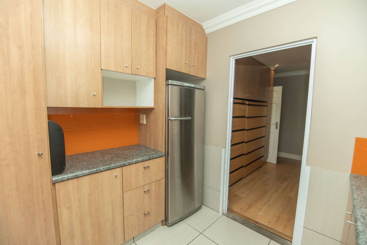 A functional kitchen area featuring light wood cabinetry, granite countertops, and a vibrant orange glass splashback, leading into an adjacent storage or utility room.