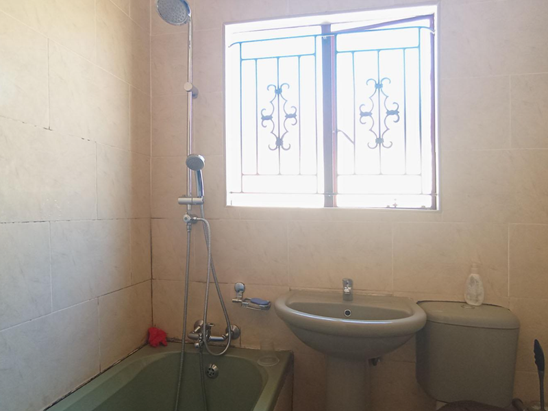 A functional bathroom featuring a bathtub with shower attachment, pedestal basin, and toilet, finished with neutral wall tiles and security bars on the window.