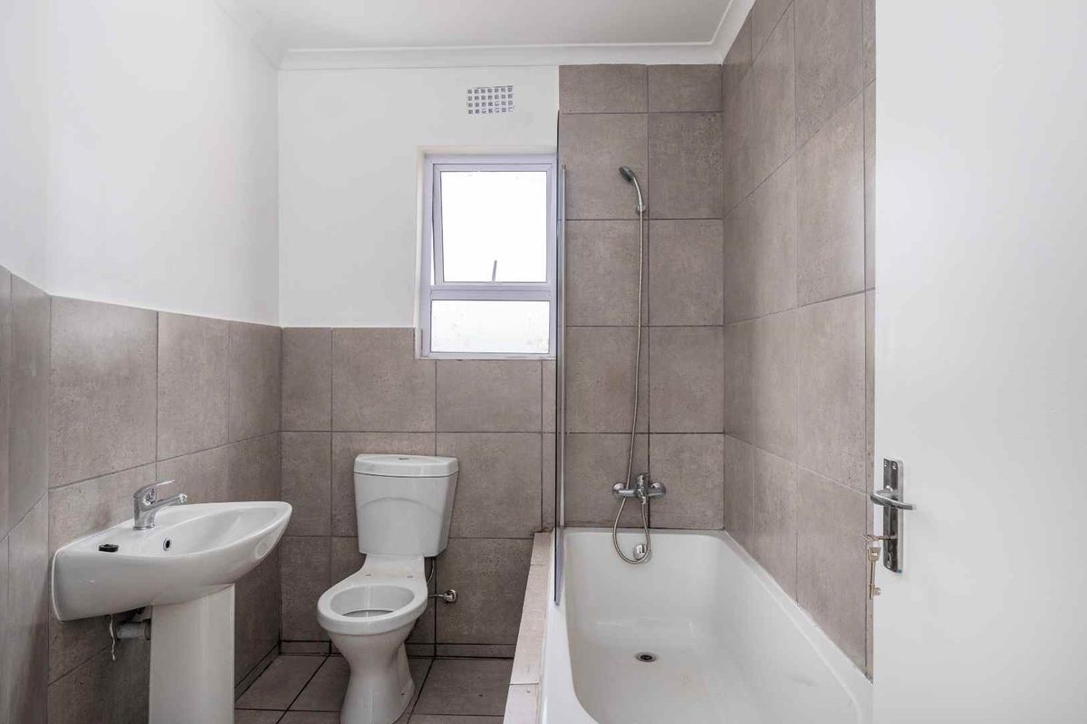 A functional bathroom featuring a pedestal basin, a standard toilet, and a shower-over-bath combination with neutral grey wall tiling.