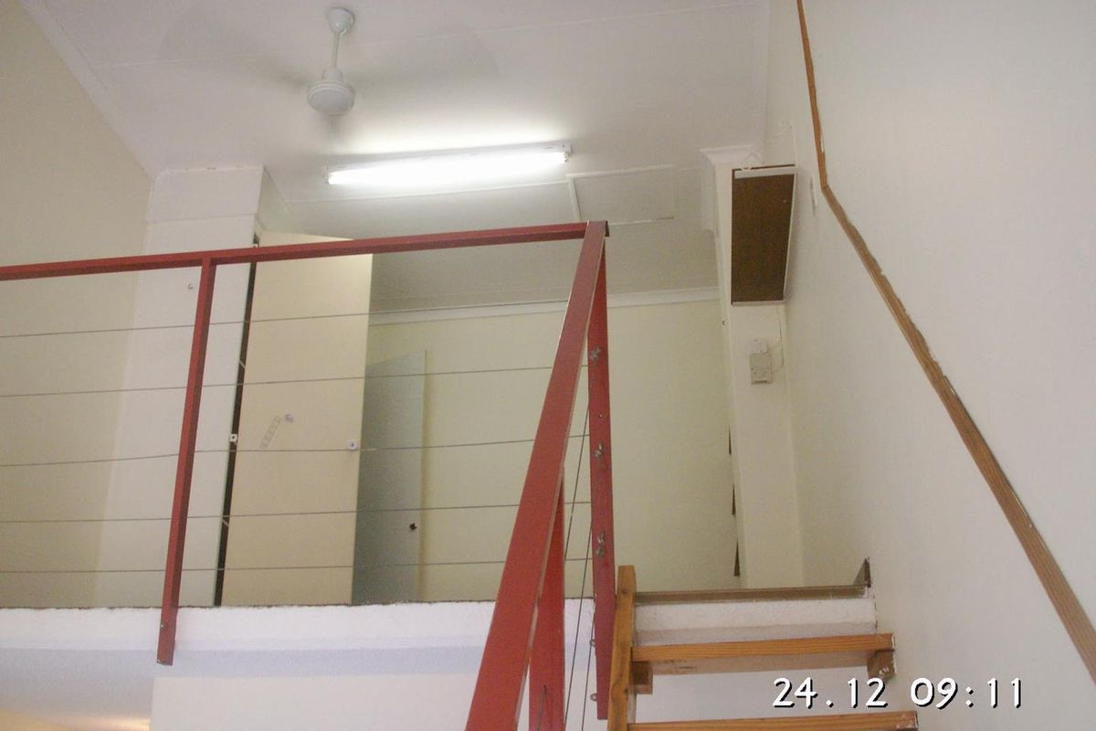 A mezzanine level bedroom area featuring a metal balustrade, timber staircase, and basic lighting fixtures, currently in need of cosmetic updates.