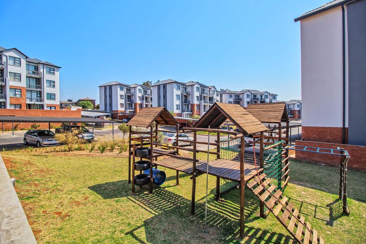 Communal outdoor area featuring a wooden playground structure set within a landscaped lawn, adjacent to apartment complex parking and residential buildings.