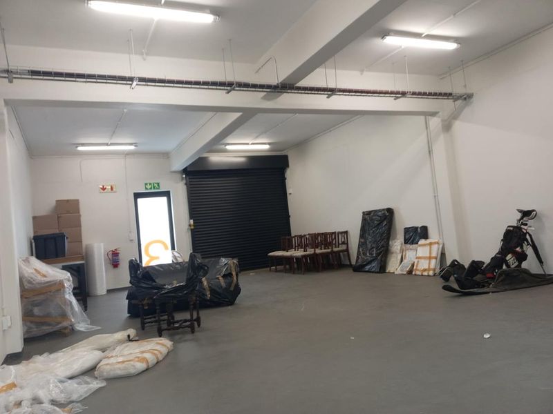 A spacious industrial utility area featuring a roller shutter door, exposed cable management, and functional overhead fluorescent lighting, suitable for storage or workshop use.