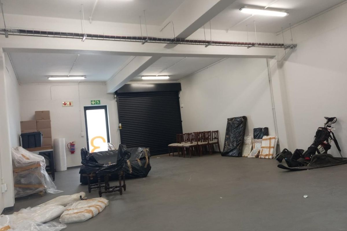 A spacious industrial utility area featuring a roller shutter door, exposed cable management, and functional overhead fluorescent lighting, suitable for storage or workshop use.
