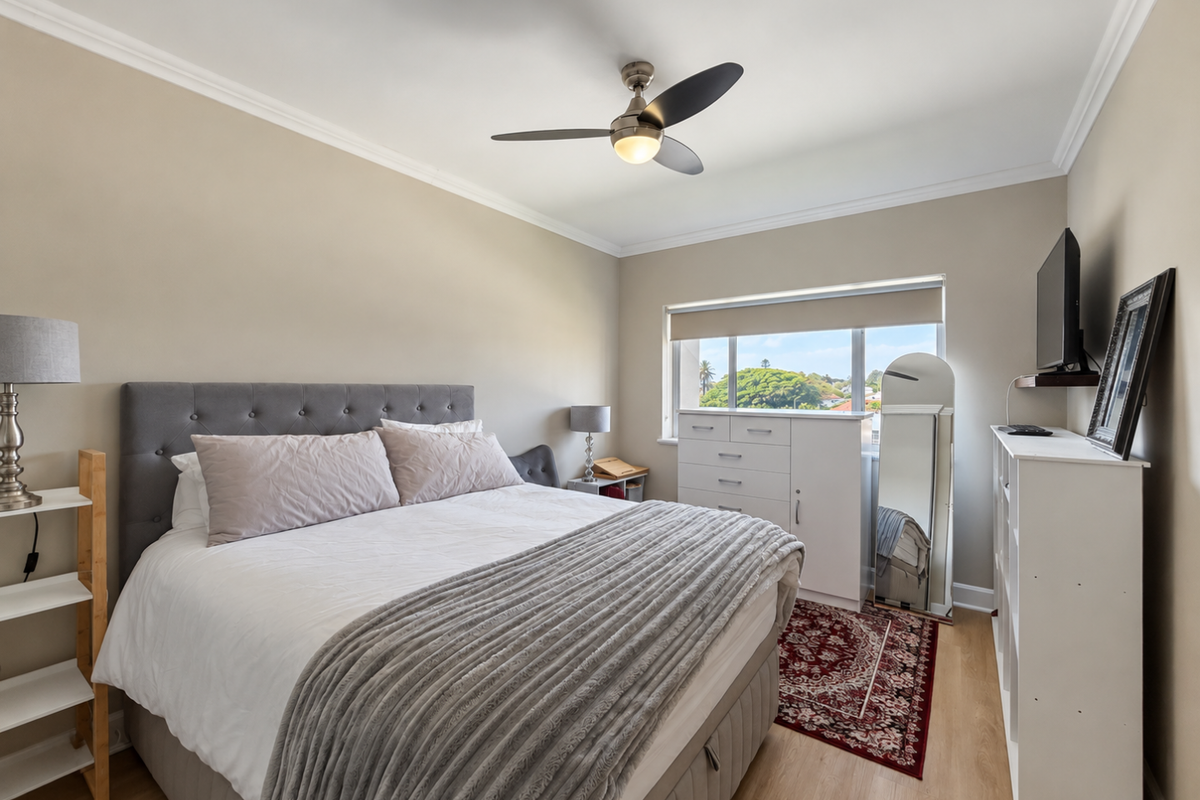 A well-maintained bedroom featuring laminate flooring, crown molding, and a ceiling fan, with natural light provided by a window overlooking the neighborhood.