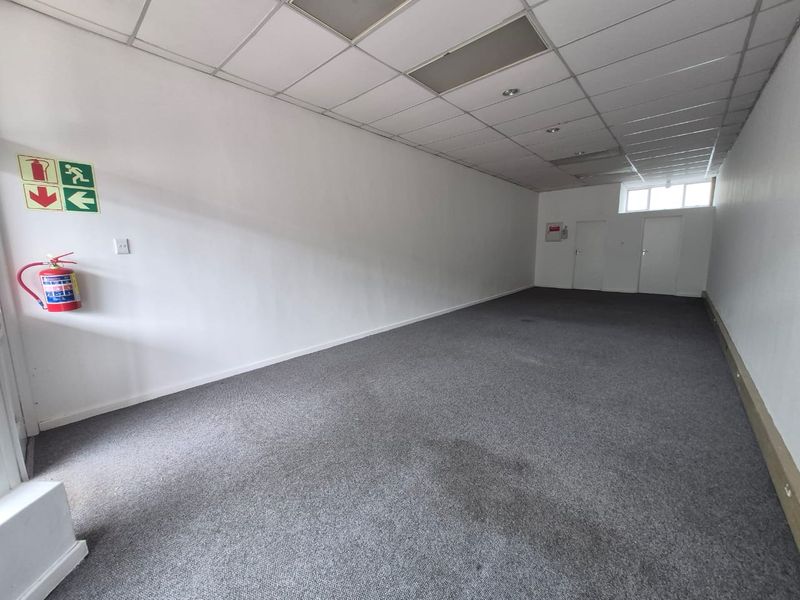 An empty, open-plan retail space featuring a suspended ceiling, recessed lighting, and carpeted flooring, equipped with essential fire safety fixtures.