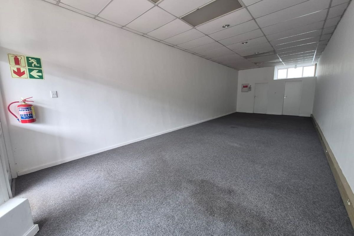 An empty, open-plan retail space featuring a suspended ceiling, recessed lighting, and carpeted flooring, equipped with essential fire safety fixtures.