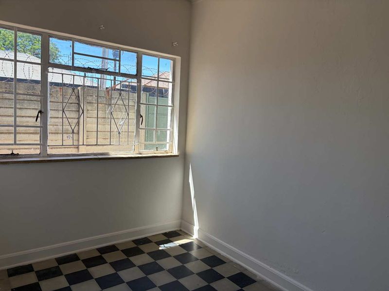 An empty bedroom featuring classic checkerboard floor tiles, a steel-framed window with security bars, and neutral walls, offering a blank canvas for renovation.