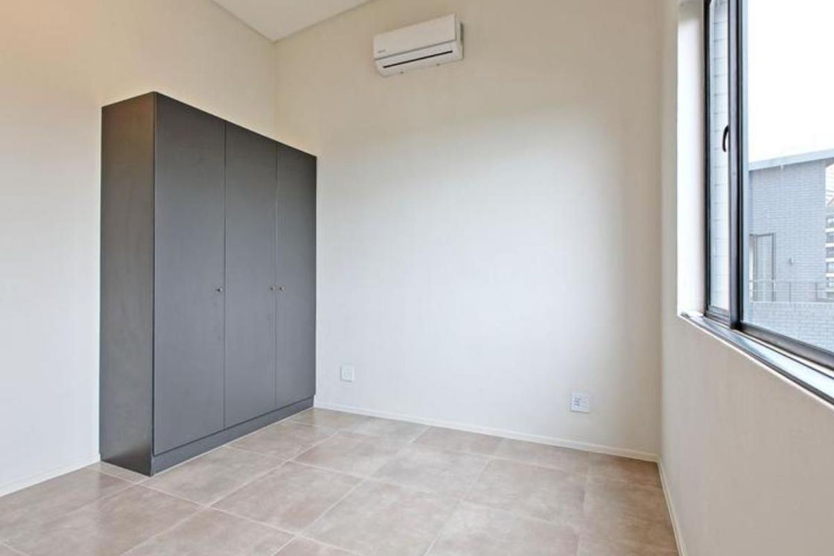 A bright, empty bedroom featuring neutral tiled flooring, a built-in dark grey wardrobe, and a wall-mounted air conditioning unit.