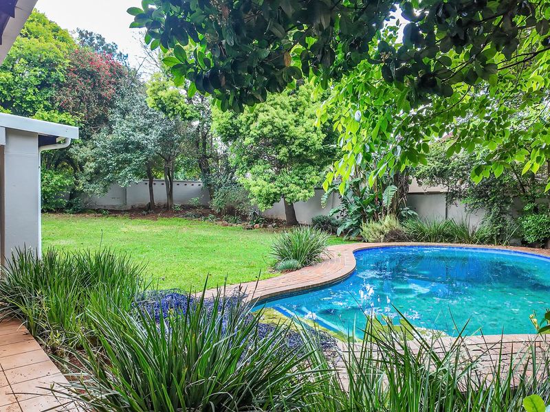 A private backyard featuring a swimming pool with brick coping, a covered patio area, and established garden landscaping.