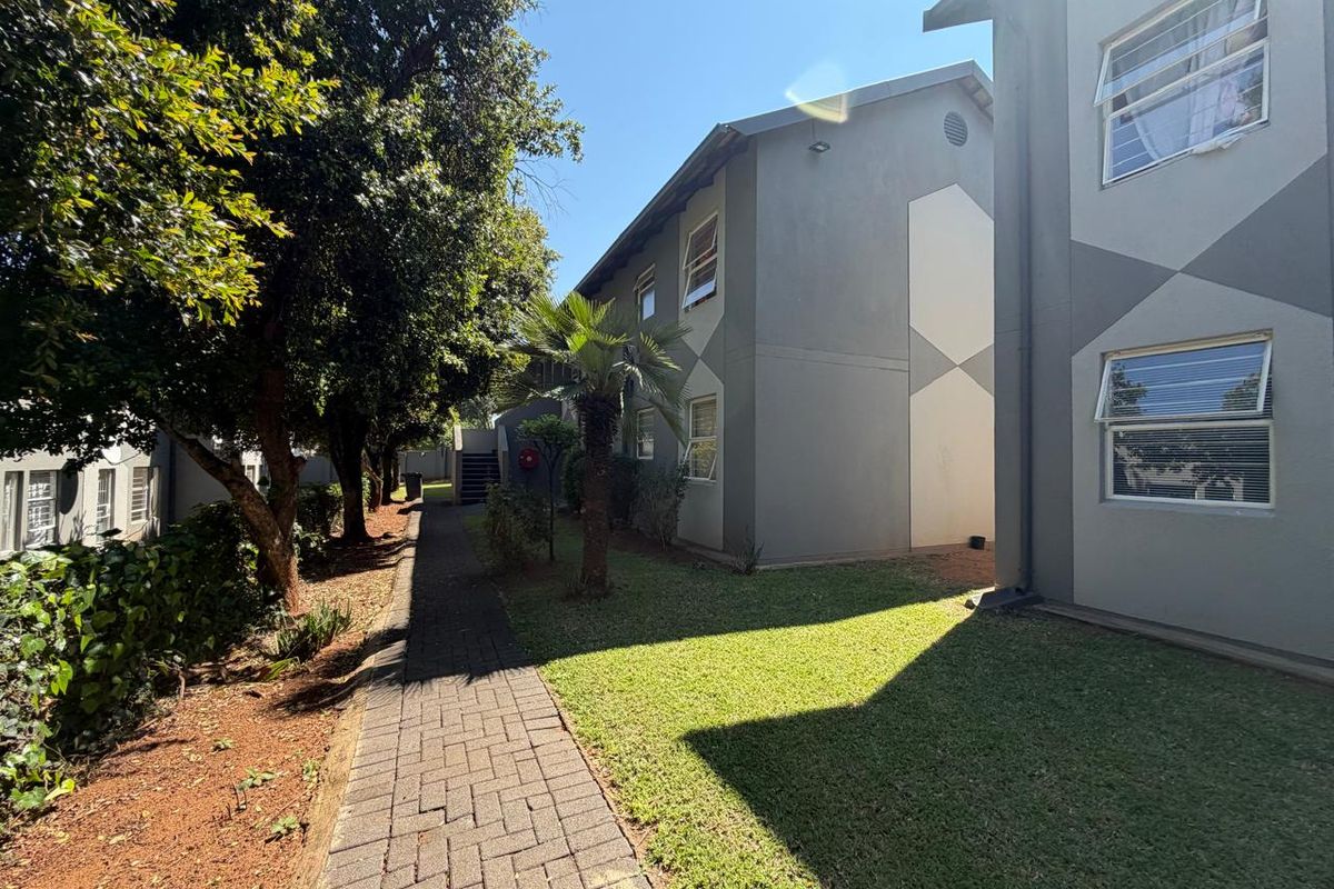 A view of the townhouse complex exterior featuring a paved walkway, well-maintained lawn, and established trees alongside the building facade.